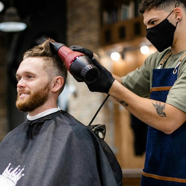 Barbier f&ouml;hnt einem l&auml;chelnden b&auml;rtigen Kunden die Haare, der Kunde tr&auml;gt eine Sch&uuml;rze