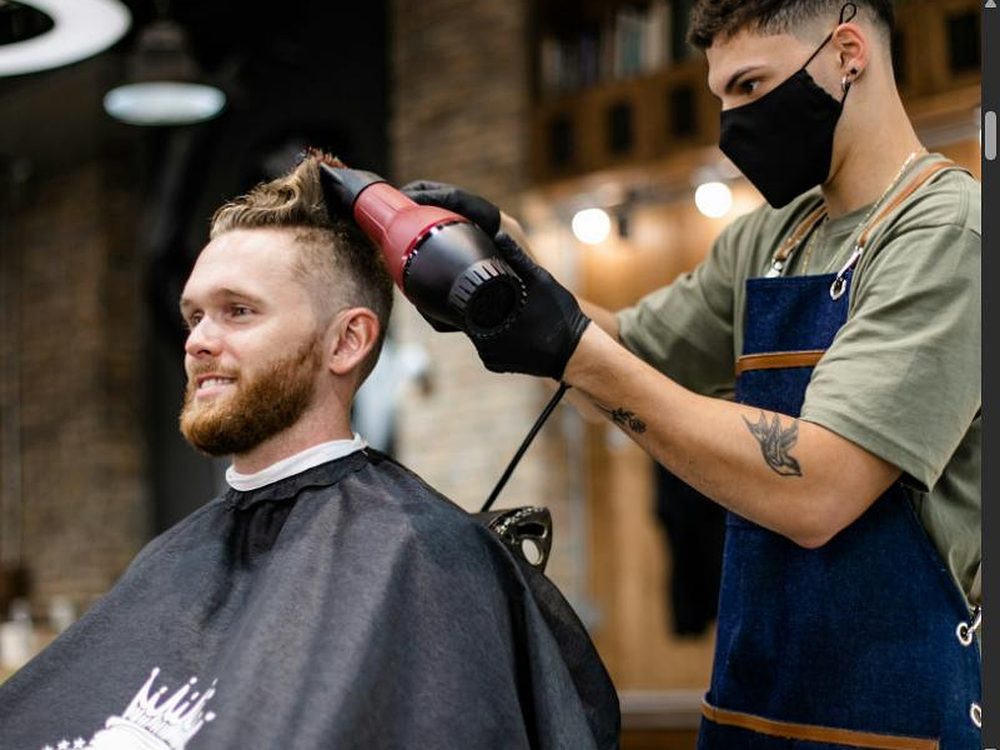 Barbier f&ouml;hnt einem l&auml;chelnden b&auml;rtigen Kunden die Haare, der Kunde tr&auml;gt eine Sch&uuml;rze