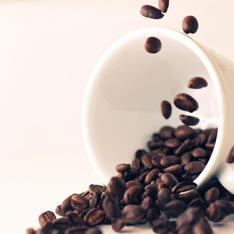 Umgekippte weiße Tasse, aus der geröstete Kaffeebohnen herausfallen, vor hellem cremefarbenem Hintergrund