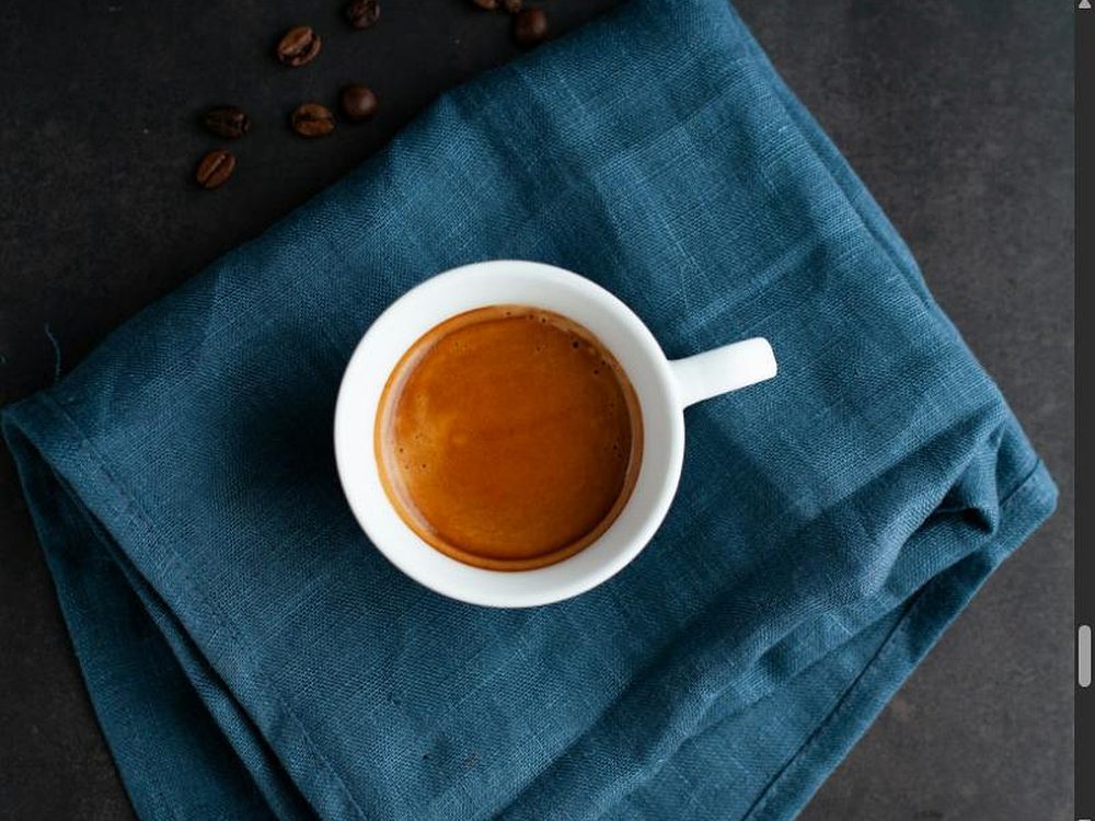 Espresso in weißer Tasse von oben auf einem petrolblauen Leinentuch, daneben einzelne Kaffeebohnen auf dunklem Steinuntergrund