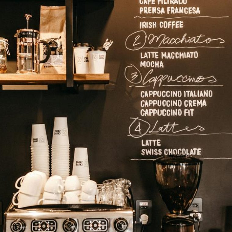 Große schwarze Tafel-Getränkekarte mit handgeschriebenen Kaffeespezialitäten, davor ein Stapel Tassen und eine Espressomaschine