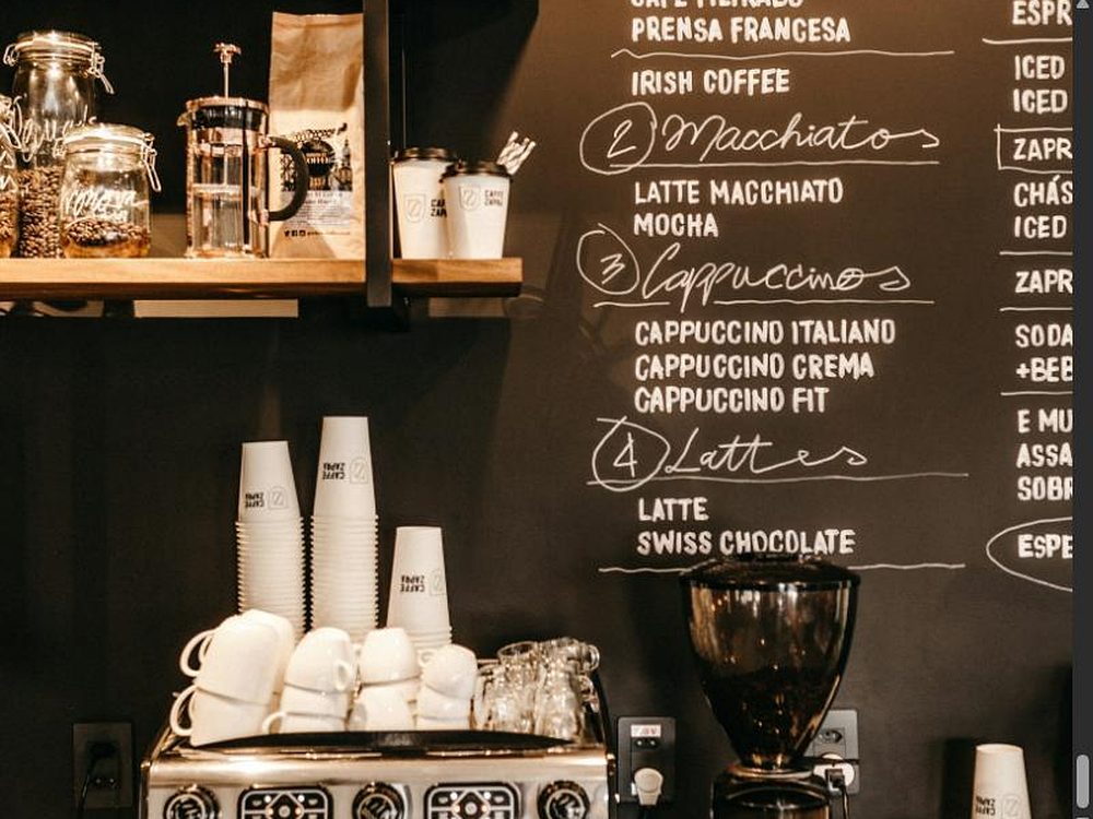Große schwarze Tafel-Getränkekarte mit handgeschriebenen Kaffeespezialitäten, davor ein Stapel Tassen und eine Espressomaschine