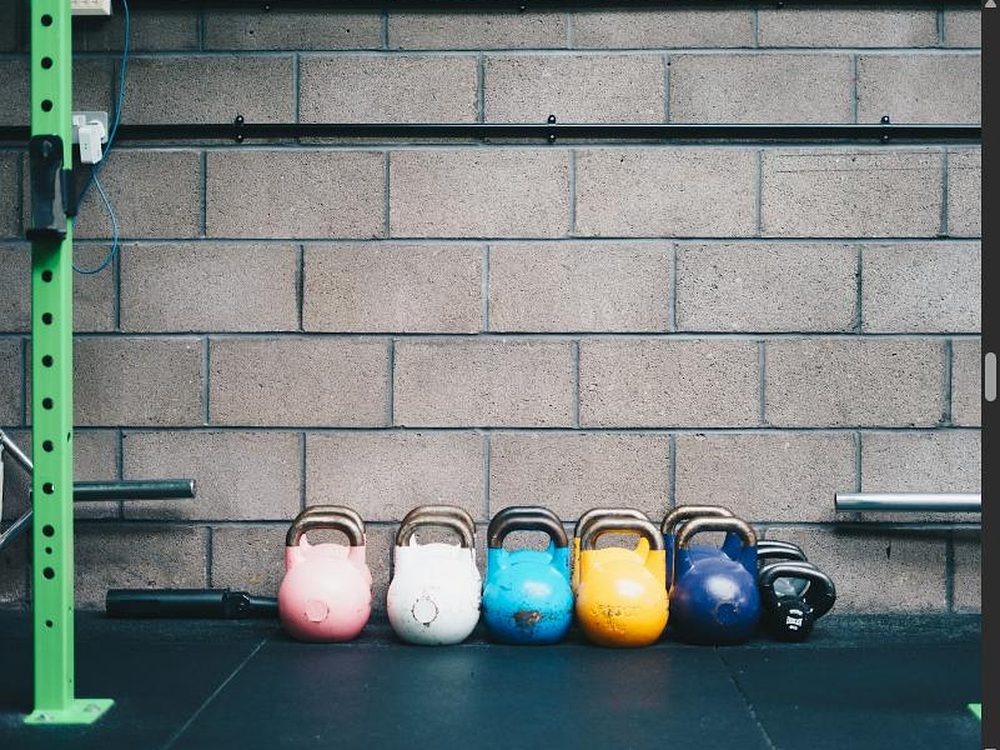 Reihe bunter Kettlebells vor einer Betonwand, daneben ein grüner Rack-Ständer im industriellen Look