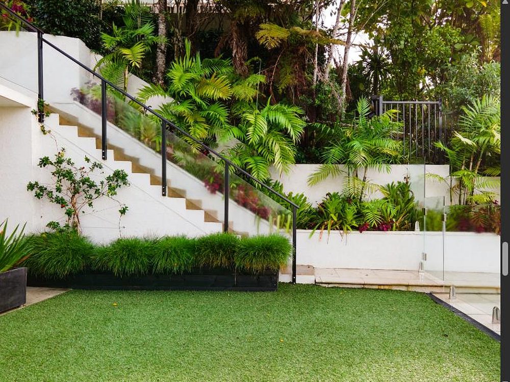 Moderne Gartenterrasse mit weißer Treppe und Glasgeländer, angrenzender Rasenfläche und dichter grüner Bepflanzung