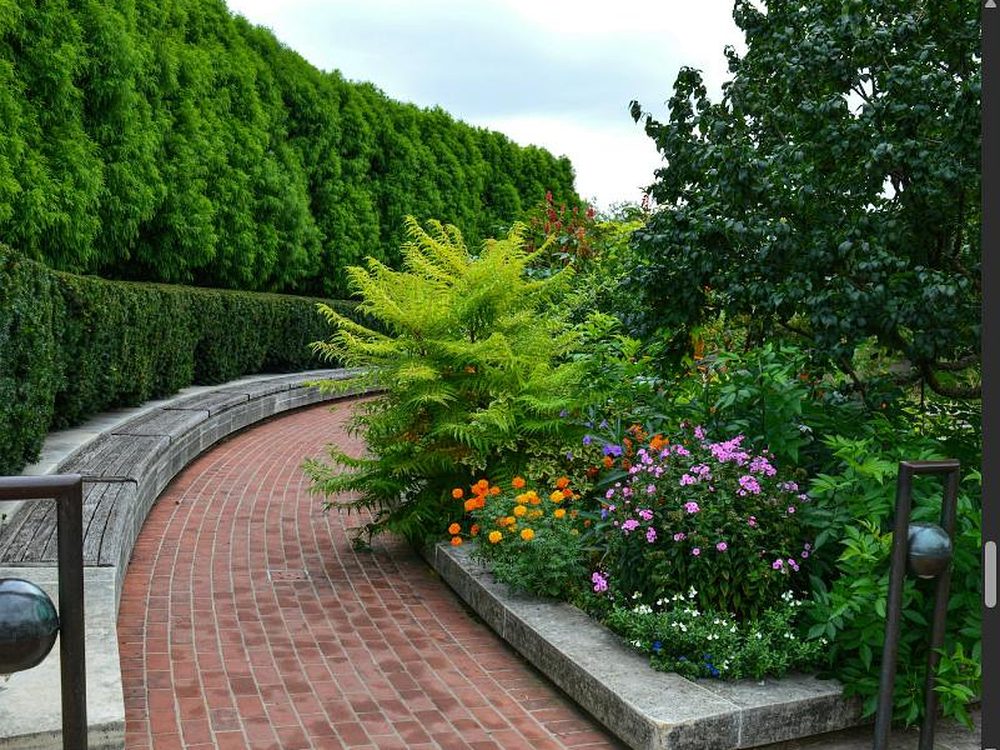 Geschwungener Klinkerweg entlang eines bunt bepflanzten Staudenbeets mit hoher Hecke und Sitzbank