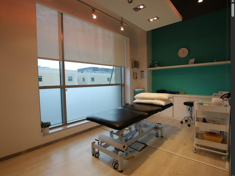 Heller Behandlungsraum mit schwarzer Therapieliege am Fenster, grüner Akzentwand, Rollhocker und Materialwagen