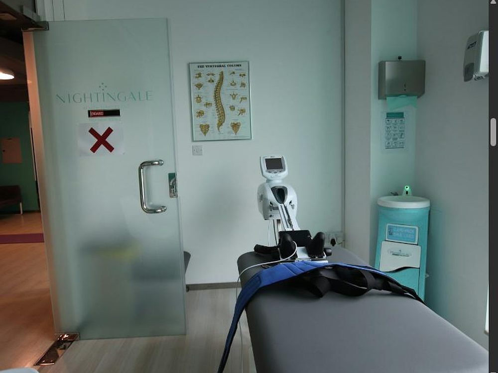 Behandlungskabine mit Therapiegerät und Gurten auf der Liege, Wirbelsäulen-Poster an der Wand und Milchglastür