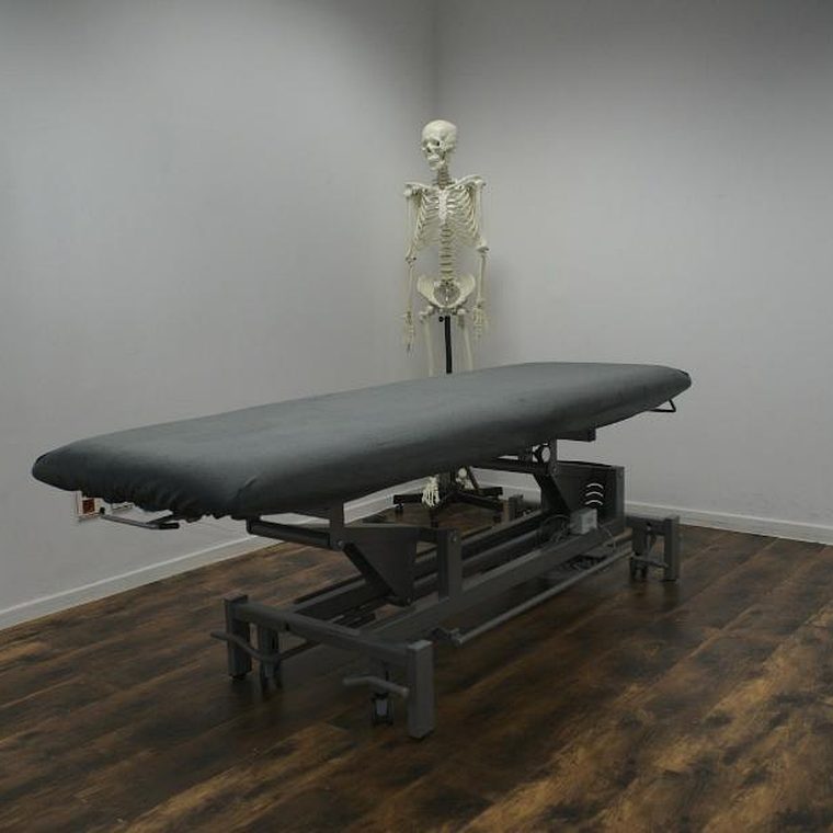 Höhenverstellbare graue Therapieliege in einem Raum mit dunklem Holzboden, dahinter an der Wand ein anatomisches Skelettmodell