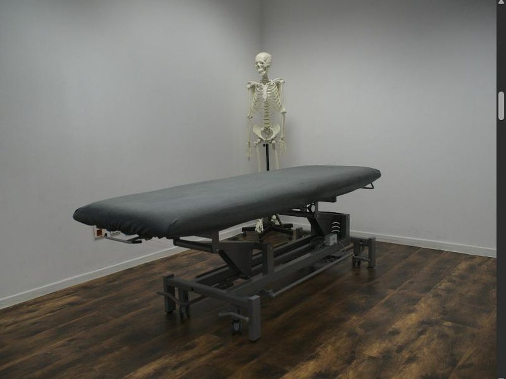 Höhenverstellbare graue Therapieliege in einem Raum mit dunklem Holzboden, dahinter ein anatomisches Skelettmodell an der Wand